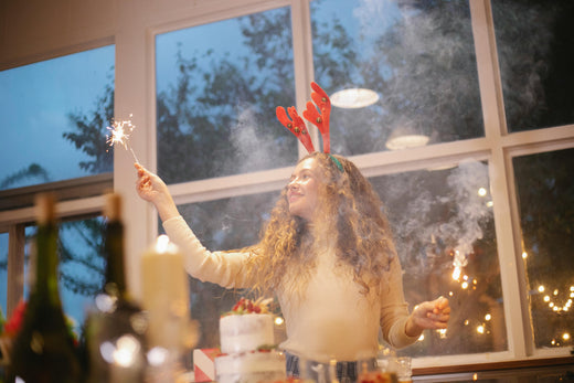 Alt Text: A joyful woman wearing reindeer antlers, holding a sparkler indoors with a festive atmosphere and twilight visible through large windows.